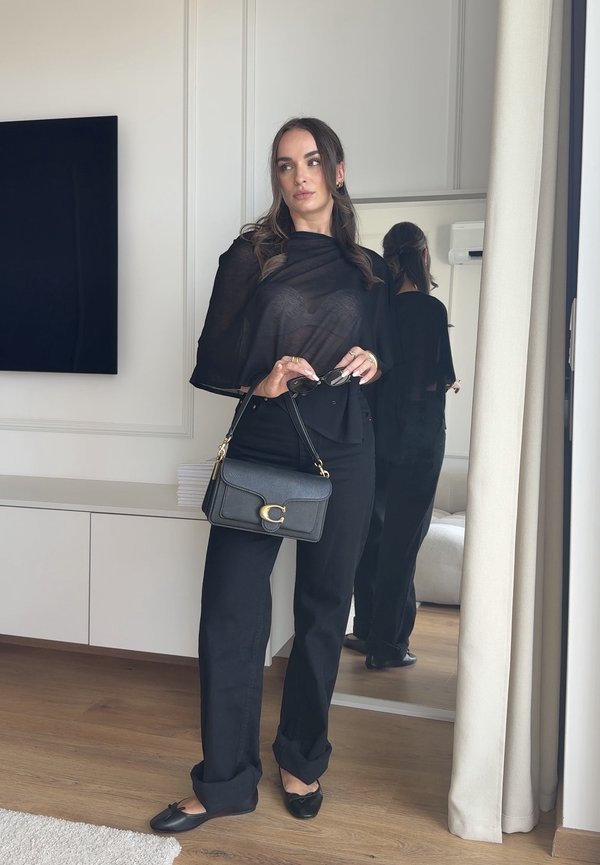 Frau trägt schwarzen transparenten Oberteil und Hose, hält schwarze Handtasche und Sonnenbrille, steht neben Spiegel in modernem Zimmer mit Holzboden.
