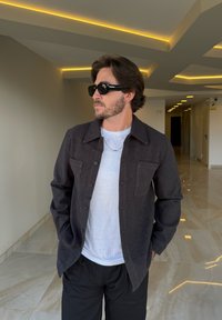 Camisa botão cinza escuro com dois bolsos frontais, usada sobre uma camiseta branca. Óculos de sol pretos e um colar de prata completam o visual.