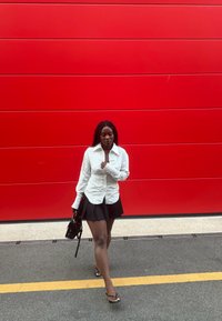Camicia a righe bianche e nere con bottoni, gonna plissettata nera, borsa nera, infradito, in piedi contro un muro rosso vivo.