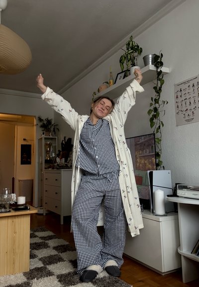 Persona estirando los brazos hacia arriba, vistiendo pijamas a cuadros y bata, de pie en una sala de estar acogedora con plantas, televisor y estanterías.