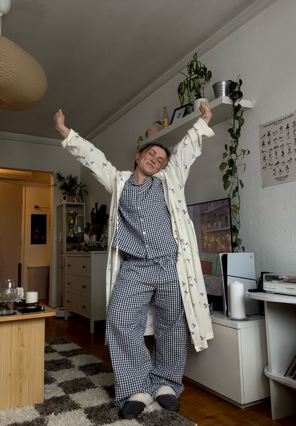 Person strækker armene op, iført ternet pyjamas og badekåbe, stående i en hyggelig stue med planter, TV og hylder.