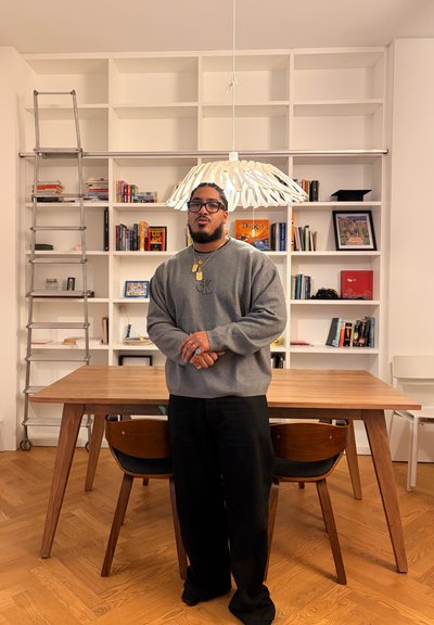 Hombre con gafas que lleva un suéter gris y pantalones negros, de pie frente a una mesa de madera y una estantería blanca con libros y una escalera.