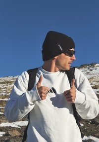 Homme en sweat-shirt blanc, bonnet noir et lunettes de soleil faisant un pouce levé en plein air sur une montagne enneigée sous un ciel bleu clair.
