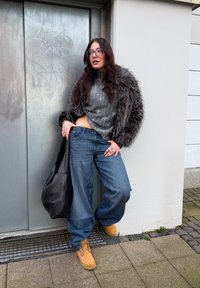 Modèle portant un pull gris côtelé, un jean en denim bleu oversize et des bottes beiges. Un sac noir repose à ses côtés, accompagné d'une veste bordée de fourrure.