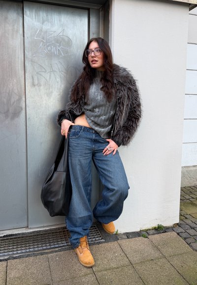 Modelo con un suéter gris acanalado, jeans azules de mezclilla oversize y botas beige. Lleva una bolsa negra a su lado y una chaqueta con ribete de piel.