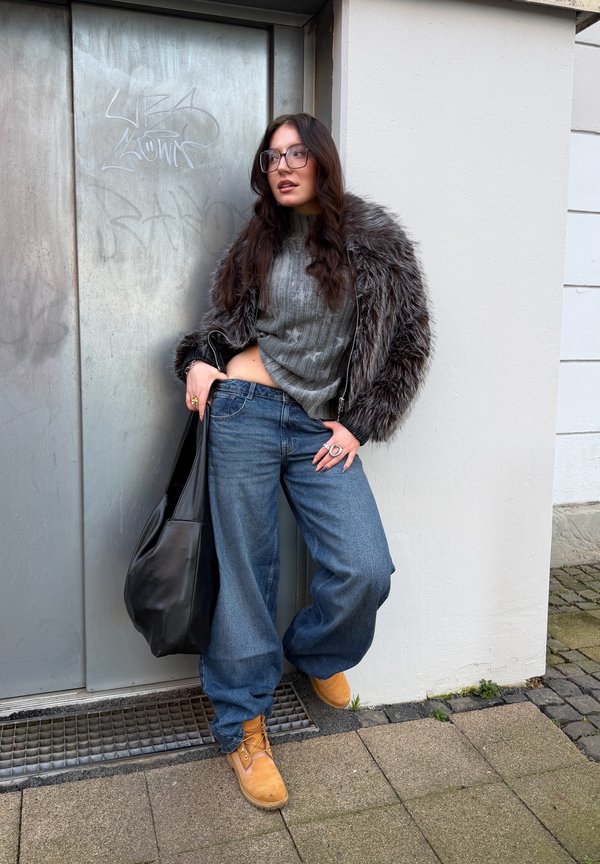 Model wearing a grey ribbed jumper, oversized blue denim jeans, and beige boots. A black bag rests at her side, with a fur-trimmed jacket.