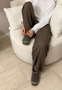 Persona seduta a gambe incrociate su un divano bianco con texture, indossa pantaloni marroni, una camicia bianca a maniche lunghe e pantofole grigie in pile, mentre sistema una pantofola.