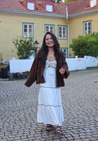Bruine koordjas over een witte maxi-jurk met bloemrijke rand, gecombineerd met bruine sandalen. Kasseien ondergrond en kleurrijke gebouwen op de achtergrond.