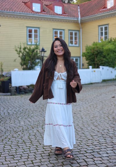 Chaqueta de pana marrón sobre un vestido maxi blanco con ribete floral, combinado con sandalias marrones. Fondo de adoquines y edificios coloridos a la vista.