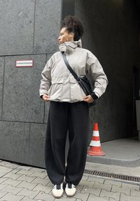 Noor curly hair wearing a light gray jacket, black wide pants, and sneakers, seis bes kõrval hall seina ja oranži liiklusmärk.