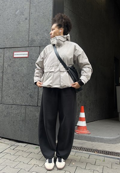 Una persona joven con cabello rizado lleva una chaqueta gris claro, pantalones anchos negros y zapatillas, de pie cerca de una pared gris y un cono de tráfico naranja.