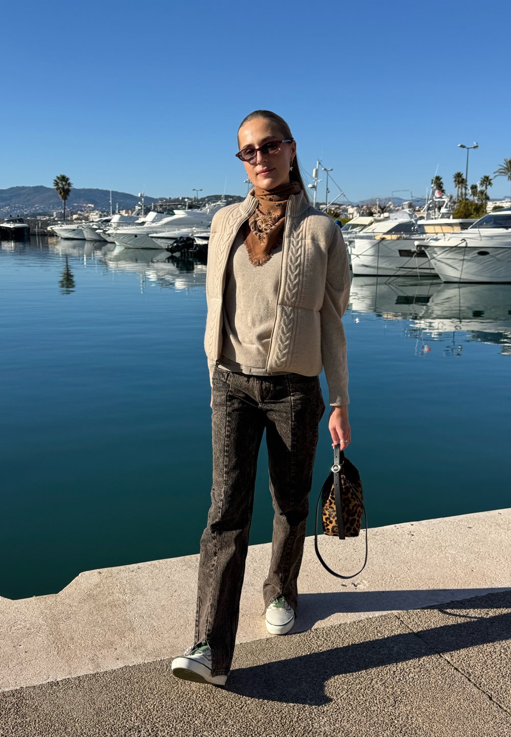 Femme en lunettes de soleil et vêtements neutres tenant un sac à imprimé léopard, marchant près d'une marina avec des yachts sous un ciel bleu clair.