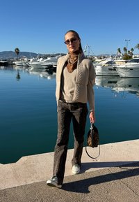 Femme en lunettes de soleil et vêtements neutres tenant un sac à imprimé léopard, marchant près d'une marina avec des yachts sous un ciel bleu clair.
