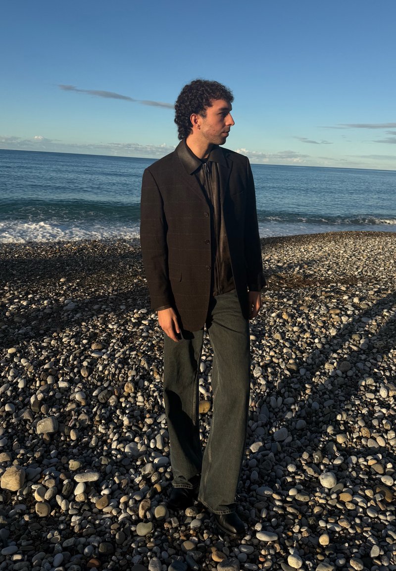Blazer a quadri scura sopra una camicia nera, abbinato a jeans di denim a gamba larga e stivali neri, con sullo sfondo una spiaggia ghiaiosa e l'oceano.