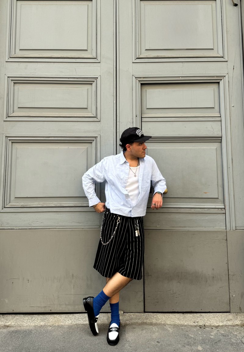 Jupe rayée noire et blanche, chemise blanche, chaussettes bleues, chaussures noires et casquette noire. Présente un détail en chaîne et des accents argentés.
