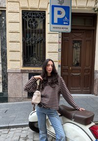 Camicia a maniche lunghe a righe marroni e bianche, jeans azzurri chiari e borsa a tracolla beige. Scooter bianco con sella marrone sullo sfondo.