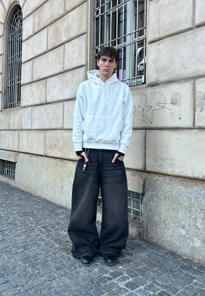 Sudadera blanca con un bolsillo frontal, combinada con jeans negros anchos y zapatos negros. Las llaves son visibles, y el fondo presenta paredes de piedra y ventanas.