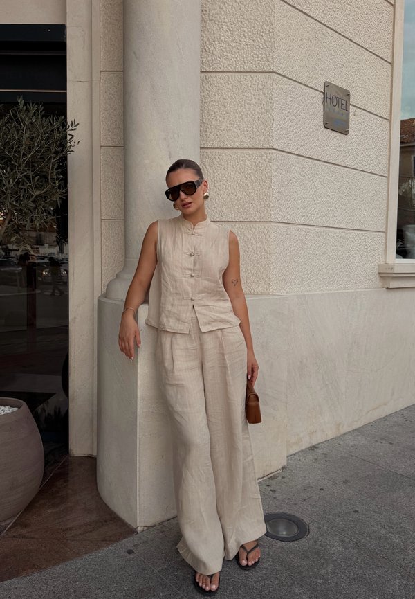Vrouw draagt een beige mouwloos topje en wijde broek, zwarte zonnebril, houdt een bruine tas vast, leunt tegen een stenen zuil buiten een gebouw met een hotelbord.