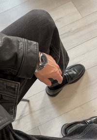 Veste en cuir noir et jeans, avec une montre-bracelet argentée dotée d'un cadran noir. Mocassins noirs à finition lisse sur un sol en bois clair.