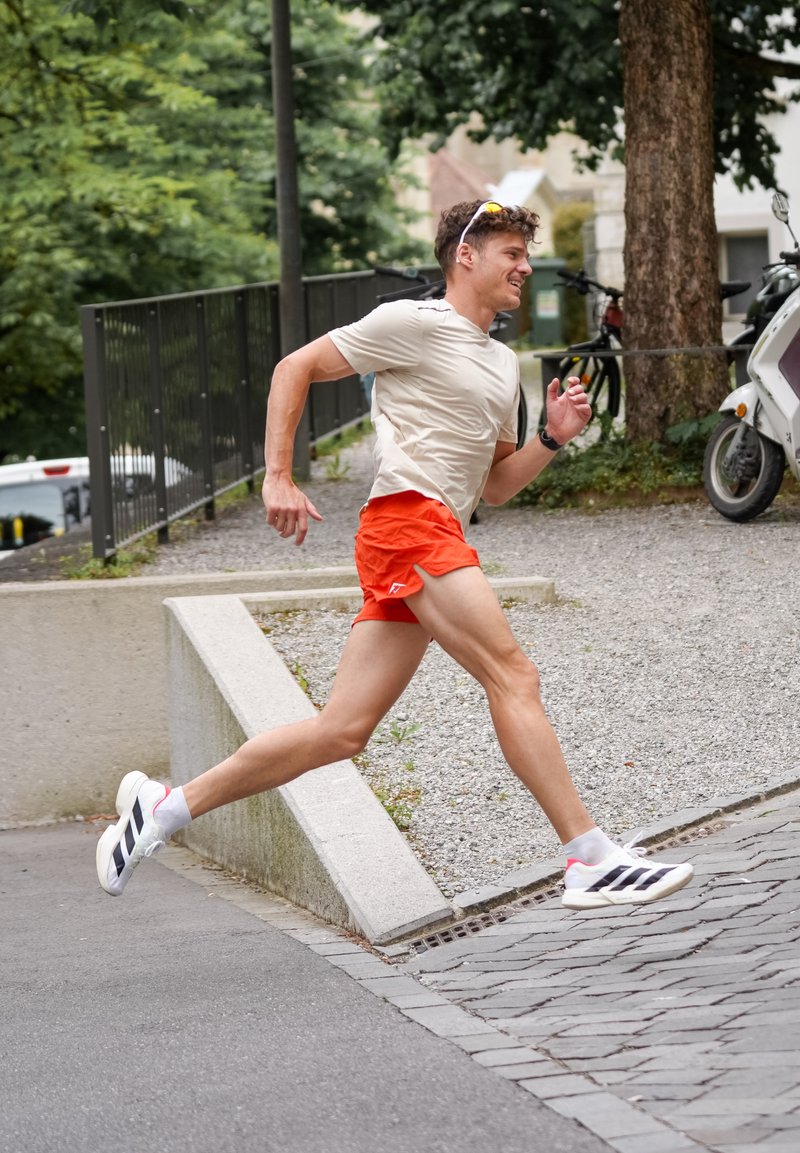 Homme en chemise claire et short rouge courant énergiquement en montée sur un trottoir en ville, entouré d'arbres, de vélos et d'une trottinette.