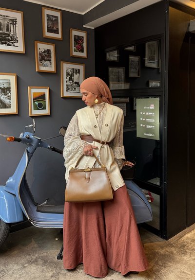 Mujer con un pañuelo beige en la cabeza y ropa de varias capas, sosteniendo un bolso marrón, apoyada en un scooter azul contra una pared oscura con fotos enmarcadas.
