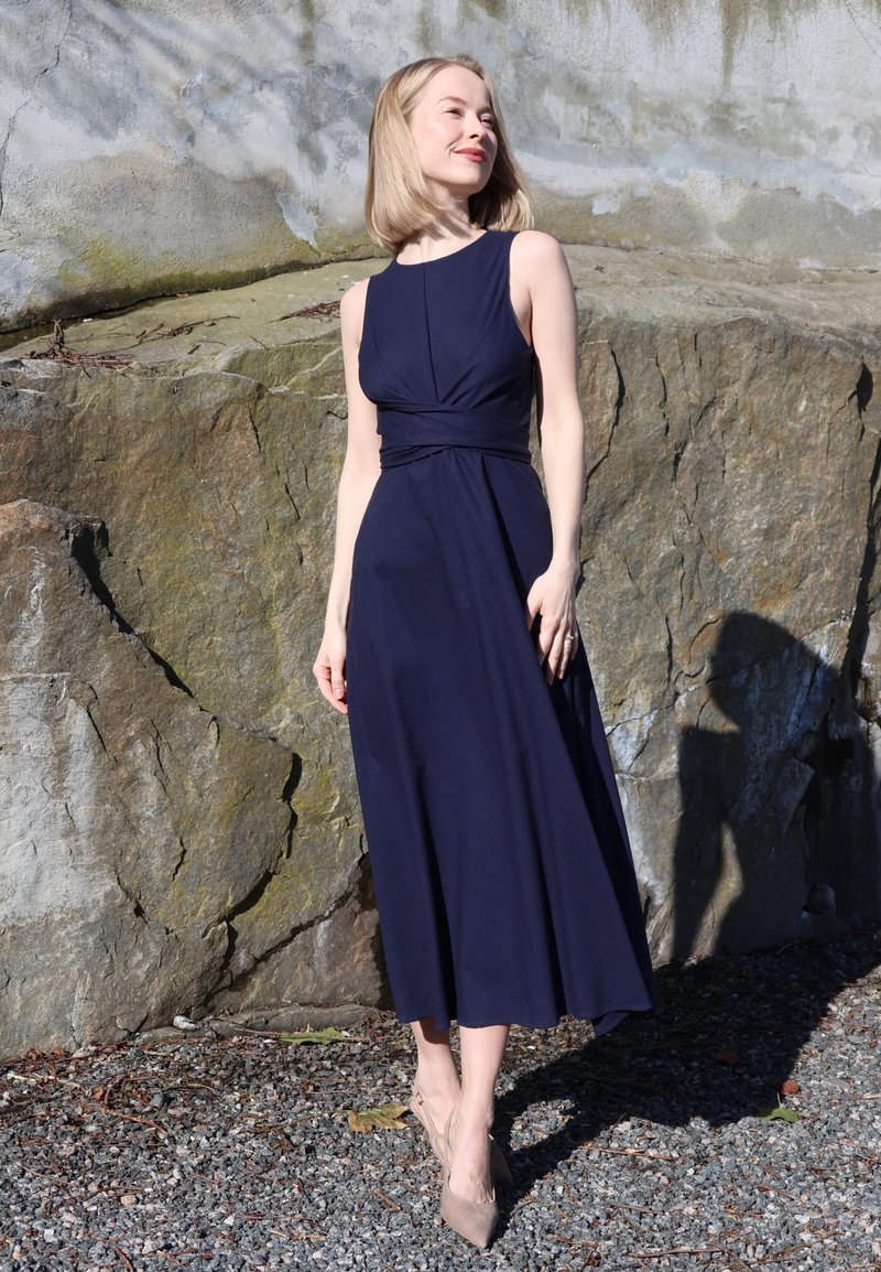 Femme en robe sans manches bleu marine et talons beige se tient sur du gravier près d'un grand mur de roche, la lumière du soleil projetant une ombre derrière elle.