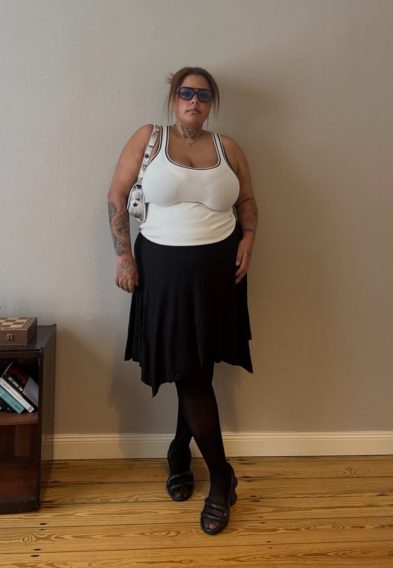 Frau mit Sonnenbrille, Tattoos, weißem Tanktop, schwarzem Rock, schwarzen Strumpfhosen und Sandalen, die an einer beigen Wand neben einem kleinen Bücherregal steht.