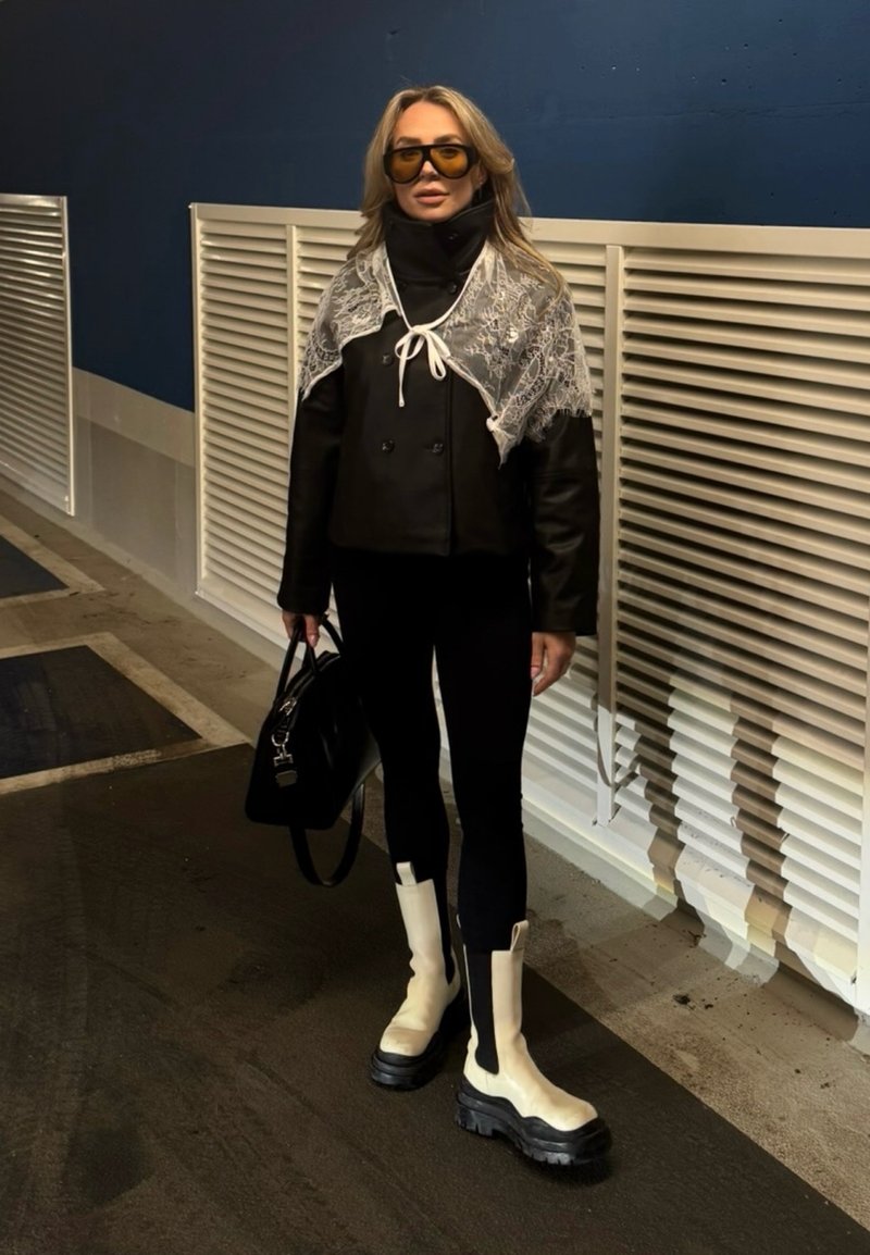 Femme portant de grandes lunettes de soleil jaunes, veste noire avec châle en dentelle, pantalon noir, bottes blanches épaisses, tenant un sac à main noir dans un cadre urbain.