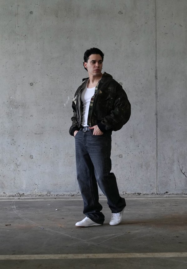 Mladý muž stojící v prostoru s betonovými stěnami, oblečený v černé bundě, bílém tílku, volných černých džínách a bílých teniskách.