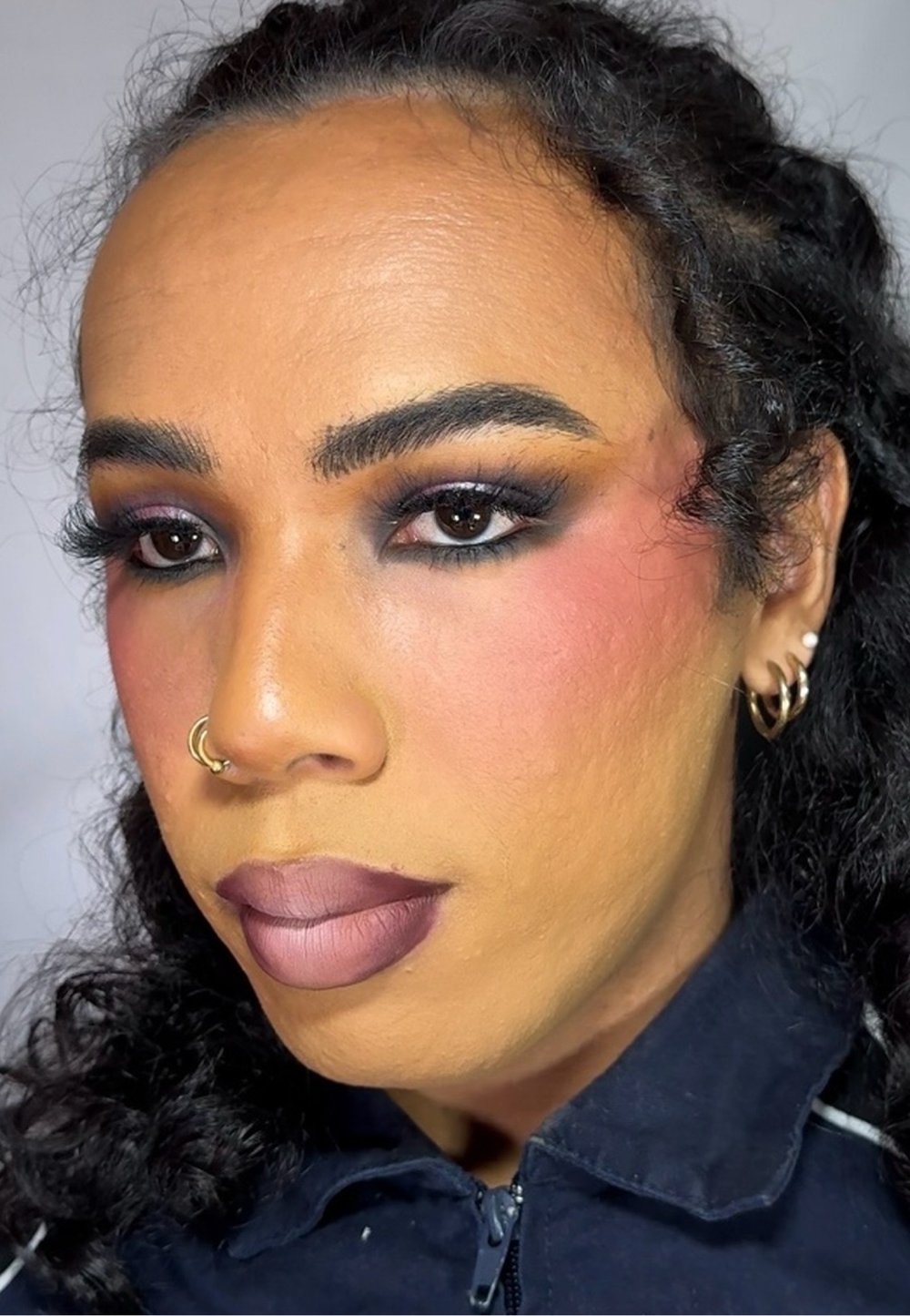 In primo piano, una persona con capelli neri ricci, che indossa una giacca scura. Ha un trucco audace: ombretto scuro, fard e labbra con una sfumatura di toni mauve.