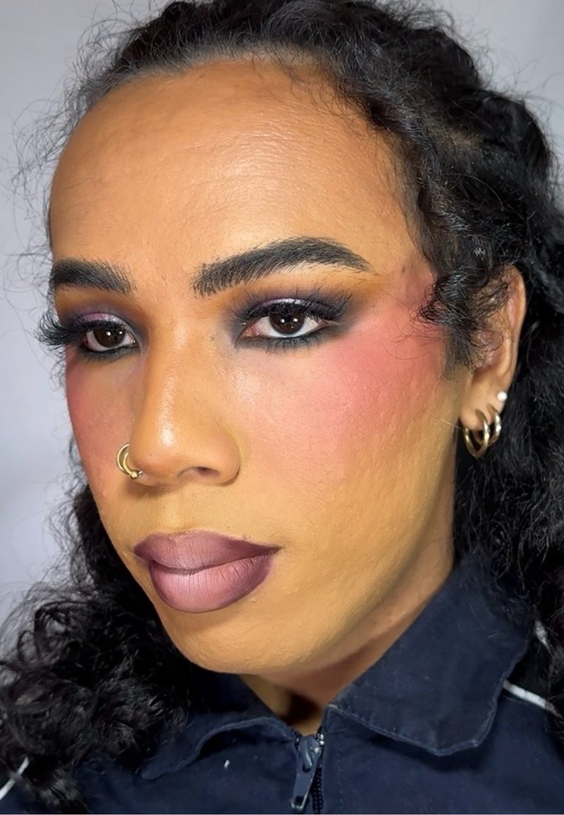 Närbild av en person med lockigt svart hår, iförd en mörk jacka. Utseendet pryds av djärv makeup: mörk ögonskugga, rouge och en gradient läpp i mauve-toner.