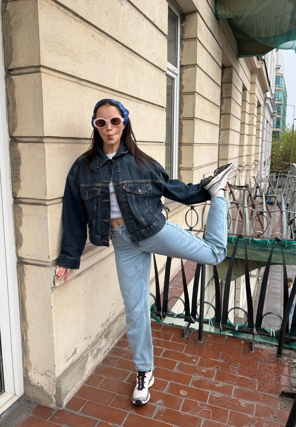 Woman in sunglasses and denim jacket stretching one leg behind on a balcony with beige stone walls and black railing.