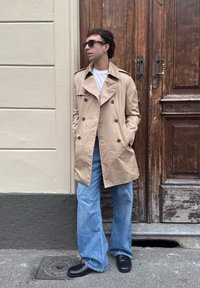 Cappotto trench beige con chiusura a doppio petto, indossato sopra una t-shirt bianca. Abbinato a jeans blu a gamba larga e scarpe nere.