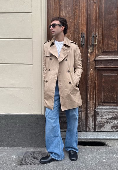 Trench beige con cierre de botón de doble botonadura, usado sobre una camiseta blanca. Combinado con jeans de pierna ancha azul y zapatos negros.
