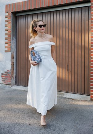 Vestido largo blanco off-the-shoulder con una silueta fluida, combinado con un bolso azul brillante y tacones nude, frente a una puerta de garaje marrón.