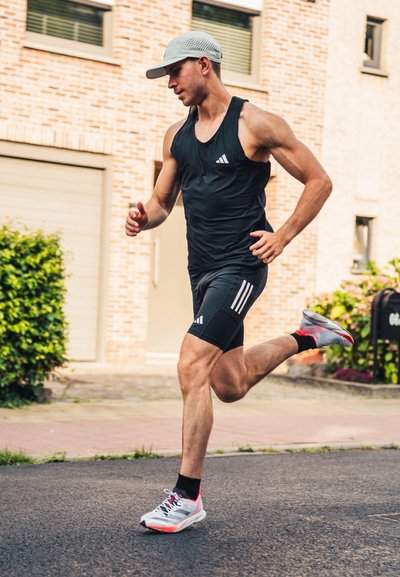 Corredor con camiseta sin mangas y pantalones cortos negros de Adidas. Gorro gris, calcetines negros y zapatillas de correr multicolores. El fondo incluye vegetación y edificios.