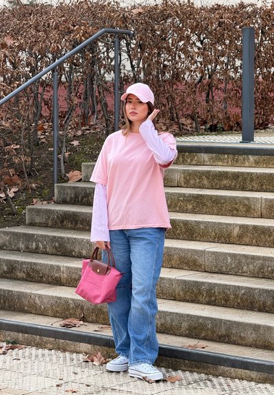 Camiseta oversized rosa claro superpuesta a una camiseta de manga larga a rayas, combinada con jeans azules descoloridos y zapatillas blancas. Sosteniendo un pequeño bolso rosa.
