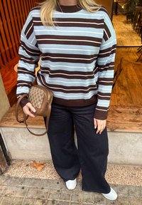 Personne portant un pull à rayures marron et bleu clair, un pantalon large noir, des chaussures blanches, tenant un sac à main marron à motifs, debout près d'un banc en bois et d'un sol en bois.