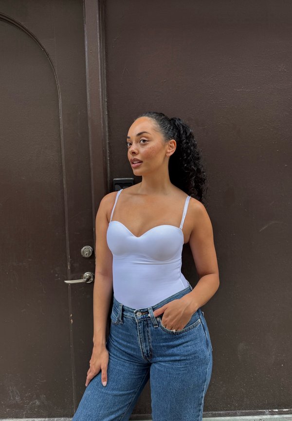 Woman with curly hair in a white tank top and blue jeans stands against a dark brown door with one hand in her pocket.