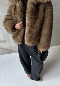 Barna műszőrme kabát széles gallérral, laza fekete nadrággal párosítva. Kiegészítők között egy kis fekete szövött clutch található. Világosszürke háttér.