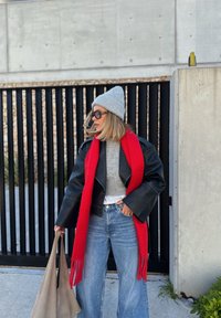 Μαύρο δερμάτινο μπουφάν, γκρι πλεκτή σκούφια, κόκκινο κασκόλ, γκρι πουλόβερ και μπλε φαρδιά τζιν, κρατώντας μια μπεζ τσάντα, σε έναν μοντέρνο φόντο.