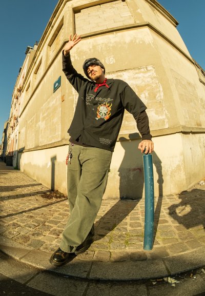 Camisa negra de botones con gráfico de llamas y calavera, pantalones verdes, zapatos negros, de pie junto a un poste azul en una calle de adoquines.