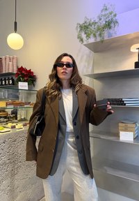 Jeune femme portant plusieurs vestes brunes superposées, une chemise blanche et un pantalon de survêtement gris, tenant un petit bol noir dans un café moderne.