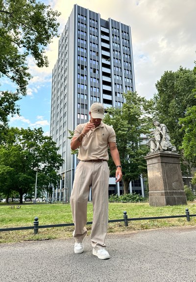 Camisa polo beige y pantalones anchos a juego, zapatillas blancas, de pie en un camino cerca de un edificio moderno alto y vegetación.