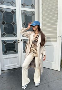Beige trench coat mintás felső felett, krémszínű, flared nadrág és ezüst sportcipő. Kék farmersapka hosszú sötét hajjal szabadtéri környezetben.