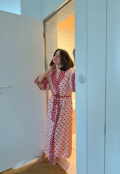 Mujer con bata estampada de pie descalza en la puerta, tocándose el cuello, con los ojos cerrados, con un baño con azulejos y luz cálida detrás de ella.