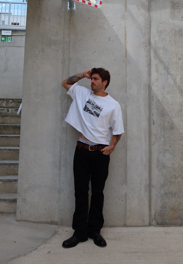 Hombre con tatuajes que lleva una camiseta blanca recortada y pantalones negros, apoyado en una pared de hormigón cerca de las escaleras, descansando la mano en la cabeza y mirando de lado.