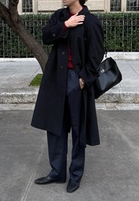 Homme portant un pardessus noir, un pantalon bleu foncé et des mocassins, debout sur un trottoir tenant un sac en cuir noir sur l'épaule.