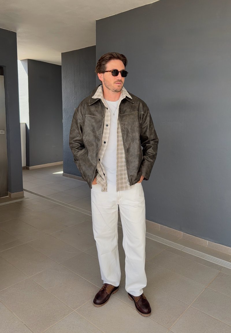 Man met zonnebril, een donkere leren jas over een geruit overhemd en witte broek, staand in een gang met grijze muren en betegelde vloer.