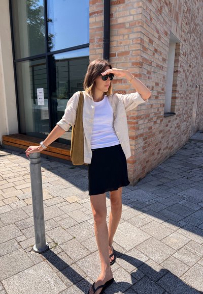 Camisa de lino beige claro sobre una camiseta blanca. Falda corta negra, chanclas negras y bolso de hombro verde oliva. Entorno urbano con pared de ladrillo.